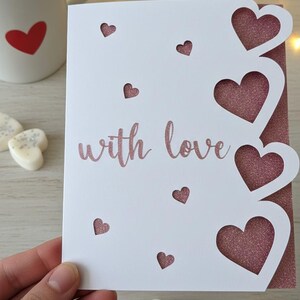 Pu&ograve; includere: Un biglietto di auguri bianco con ritagli a forma di cuore e la scritta "with love" in un carattere glitterato rosa. Il biglietto ha un retro glitterato rosa. Sullo sfondo una tazza bianca con un cuore rosso.