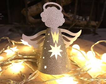 May include: Glittery silver angel Christmas ornament with a halo, wings, and a star-shaped cutout on the cone-shaped body. The angel is surrounded by a string of warm white star-shaped lights, creating a festive holiday scene.