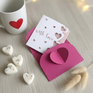 Pu&ograve; includere: Una tazza bianca con un cuore rosso, una busta rosa con un ritaglio a forma di cuore e una carta con la scritta "with love". Cuori di cera profumata e rametti decorativi completano il tema romantico.