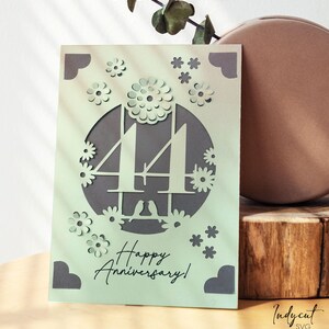 May include: A light green paper anniversary card with a cut-out design. The card features the number "44" surrounded by flowers and the words "Happy Anniversary!". The card is next to a brown vase and a wooden block.