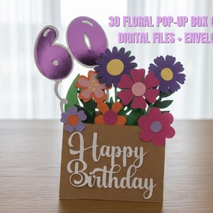 May include: A 3D floral pop-up box card with a brown box base and colorful paper flowers. The card features the number 60 in purple and the words "Happy Birthday" in white script. The text "3D Floral Pop-Up Box Card Digital Files + Envelope" is also visible.