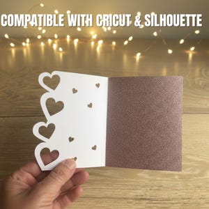Pu&ograve; includere: Un biglietto bianco con ritagli a forma di cuore e un interno marrone glitterato &egrave; tenuto aperto. Il biglietto &egrave; su una superficie di legno, con luci a corda sullo sfondo. Il testo "COMPATIBILE CON CRICUT & SILHOUETTE" &egrave; in alto.