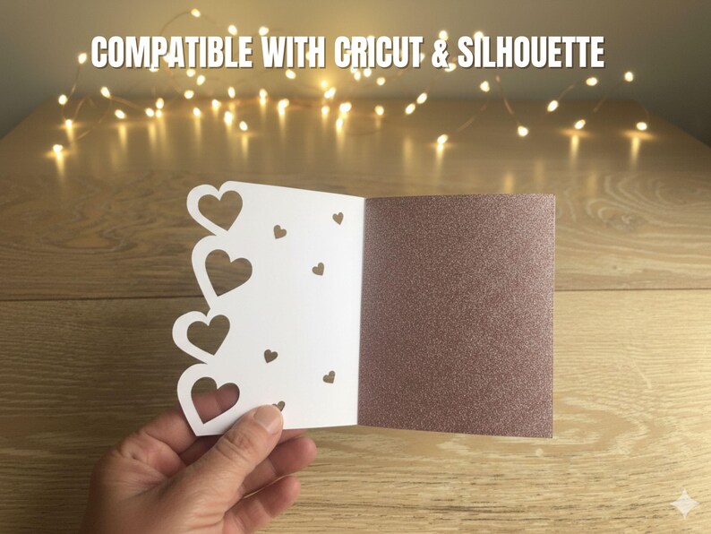 Pu&ograve; includere: Un biglietto bianco con ritagli a forma di cuore e un interno marrone glitterato &egrave; tenuto aperto. Il biglietto &egrave; su una superficie di legno, con luci a corda sullo sfondo. Il testo "COMPATIBILE CON CRICUT & SILHOUETTE" &egrave; in alto.