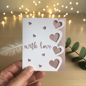 Pu&ograve; includere: Un biglietto bianco con cuori ritagliati e la scritta "with love" in una scrittura delicata. Il biglietto ha un retro rosa glitterato e piccoli ritagli a forma di cuore sono sparsi sul davanti. Una piuma e del verde sono sullo sfondo.