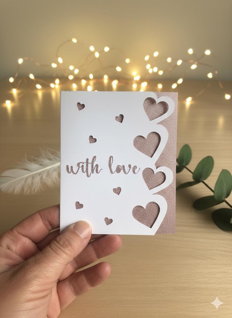Pu&ograve; includere: Un biglietto bianco con cuori ritagliati e la scritta "with love" in una scrittura delicata. Il biglietto ha un retro rosa glitterato e piccoli ritagli a forma di cuore sono sparsi sul davanti. Una piuma e del verde sono sullo sfondo.