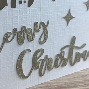 May include: White Christmas card with gold glitter lettering that reads "Merry Christmas." The card has a textured surface and features a cityscape silhouette and star-shaped accents.