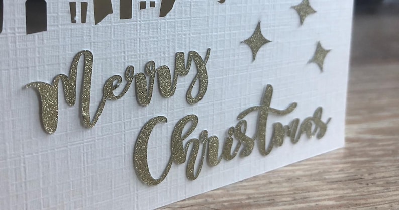 May include: White Christmas card with gold glitter lettering that reads "Merry Christmas." The card has a textured surface and features a cityscape silhouette and star-shaped accents.
