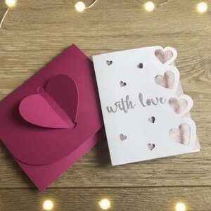 Pu&ograve; includere: Un biglietto di auguri bianco con un design a cuore glitterato rosa e le parole "with love" accanto a una busta rosa abbinata con un ritaglio a forma di cuore. Il biglietto ha ritagli a forma di cuore. Sfondo in legno con luci a corda.