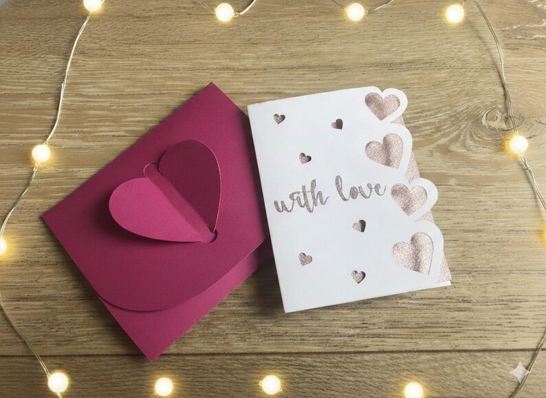 Pu&ograve; includere: Un biglietto di auguri bianco con un design a cuore glitterato rosa e le parole "with love" accanto a una busta rosa abbinata con un ritaglio a forma di cuore. Il biglietto ha ritagli a forma di cuore. Sfondo in legno con luci a corda.