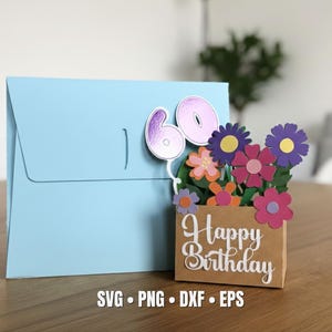 May include: A light blue birthday card with a 3D floral arrangement and the number 60. The flowers are various colors, including purple, pink, and orange. The card also has the words "Happy Birthday" on the front.