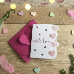 Pu&ograve; includere: Un biglietto bianco con la scritta "with love" e ritagli a forma di cuore, accanto a una busta rosa e piccoli cuori di legno. La scena &egrave; disposta su una superficie di legno con piume e rosmarino.