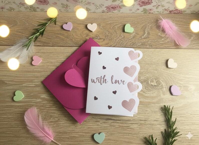 Pu&ograve; includere: Un biglietto bianco con la scritta "with love" e ritagli a forma di cuore, accanto a una busta rosa e piccoli cuori di legno. La scena &egrave; disposta su una superficie di legno con piume e rosmarino.