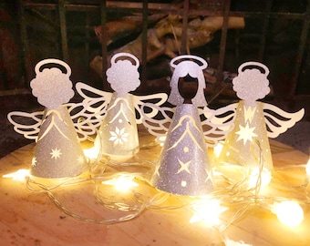 May include: Four glittery silver angel ornaments with cut-out wings and halos, illuminated by a string of warm white star-shaped lights. The angels have cone-shaped bodies with star and floral cutouts. The scene is set on a wooden surface.