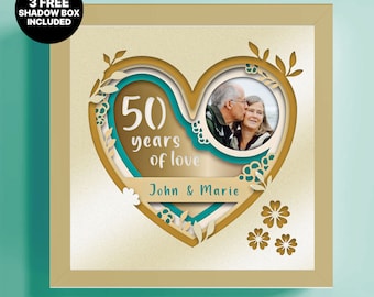 May include: A framed, layered paper art piece celebrating a 50th wedding anniversary. The design features a heart shape with the words "50 years of love" and the names "John & Marie." A photo of a couple is included. The frame is gold.