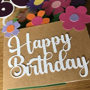 May include: A birthday card with the words "Happy Birthday" in white script. The card is decorated with colorful paper flowers in shades of pink, purple, and orange. A number 60 is also visible.