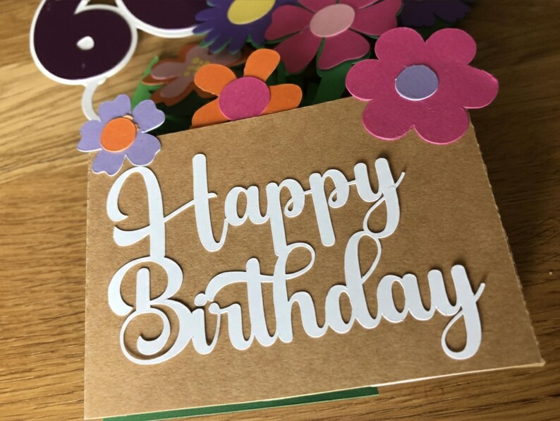 May include: A birthday card with the words "Happy Birthday" in white script. The card is decorated with colorful paper flowers in shades of pink, purple, and orange. A number 60 is also visible.