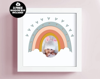 May include: A white frame with a rainbow design in shades of blue, pink, and yellow. The rainbow has a cutout in the center, revealing a photo of a baby wearing a pink and white hat. The rainbow is surrounded by white clouds and small white hearts.