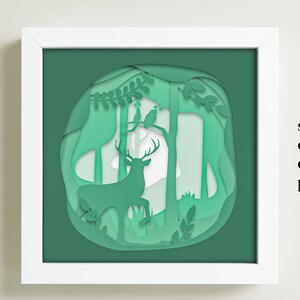 May include: A green paper cut-out design of a deer in a forest with trees and birds. The design is framed in a white frame.
