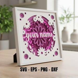 May include: A white framed, layered paper art piece with a floral design. The artwork features a central circle with the words "your name" in a script font. The design includes white daisies and purple floral elements. The frame is supported by a black stand.