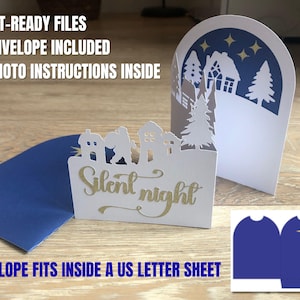 以下が含まれることがあります： 白い紙のクリスマスカード。切り抜かれた村のシーンと、金色の筆記体で書かれた「Silent night」の文字。青い封筒が含まれています。カードには、家、木、星空のある冬のシーンが描かれています。