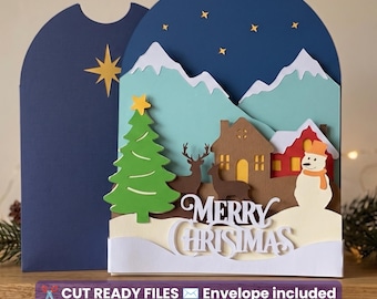 May include: A layered paper craft Christmas card featuring a dark blue night sky with yellow stars, light blue mountains, a green tree, brown deer, a red house, and a white snowman. The white text reads Merry Christmas.