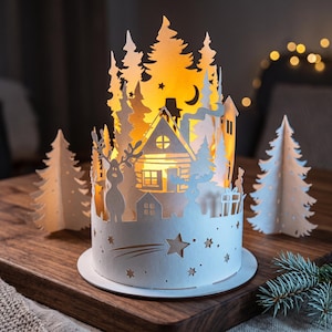 May include: A cylindrical white paper lantern featuring intricate laser-cut silhouettes of a cabin, pine trees, a deer, and a crescent moon. The structure is illuminated from within, casting a warm glow through the cutouts, with two separate paper pine trees placed on either side.