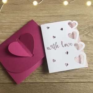 Pu&ograve; includere: Un biglietto di auguri bianco con la scritta "with love" e ritagli a forma di cuore, accanto a una busta magenta con un design a forma di cuore. Il biglietto presenta cuori rosa glitterati. Su una superficie di legno con luci a filo.