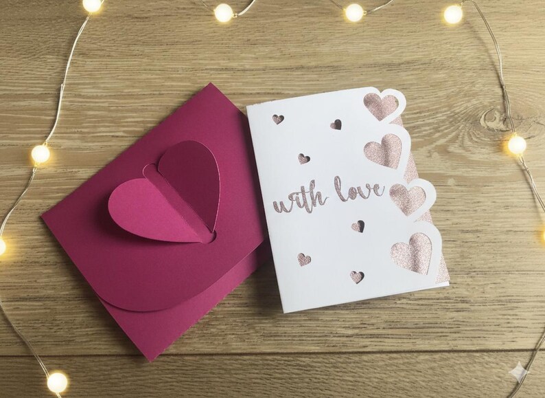 Pu&ograve; includere: Un biglietto di auguri bianco con la scritta "with love" e ritagli a forma di cuore, accanto a una busta magenta con un design a forma di cuore. Il biglietto presenta cuori rosa glitterati. Su una superficie di legno con luci a filo.