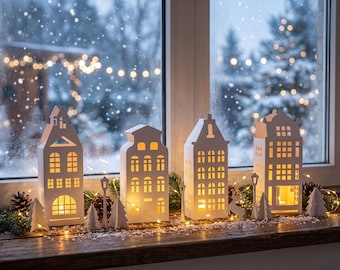 May include: Four white house-shaped lanterns with cut-out windows and doors, illuminated from within, arranged on a wooden surface with pine cones, small paper trees, a deer figure, and string lights against a snowy window background.