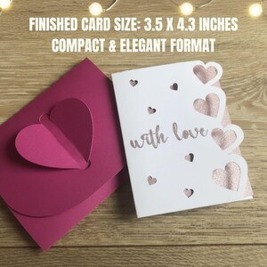 Pu&ograve; includere: Un biglietto magenta con un cuore a comparsa e un biglietto bianco con ritagli a forma di cuore e la scritta "with love". Le dimensioni finali del biglietto sono 8,9 x 10,9 cm. I biglietti sono su una superficie di legno.