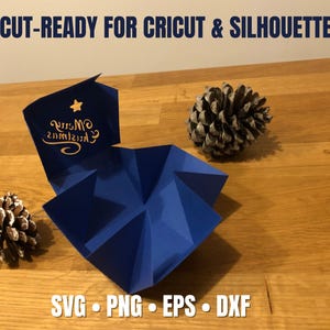 May include: A dark blue, origami-style paper box with a gold star and the words "Merry Christmas" in gold script. Two pine cones sit on a wooden surface. Text at the top reads "CUT-READY FOR CRICUT & SILHOUETTE".