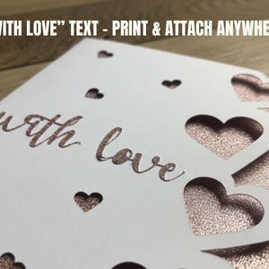 Pu&ograve; includere: Un biglietto bianco con ritagli a forma di cuore e le parole "with love" in un carattere oro rosa glitterato. Il biglietto ha un retro glitterato oro rosa. Il testo "WITH LOVE" - PRINT & ATTACH ANYWHERE &egrave; in alto.