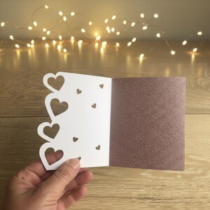 Pu&ograve; includere: Un biglietto di auguri bianco a forma di cuore, con un interno rosa dorato glitterato. Il biglietto presenta una serie di ritagli a forma di cuore e piccoli cuori decorativi. Il biglietto &egrave; aperto, tenuto in mano.