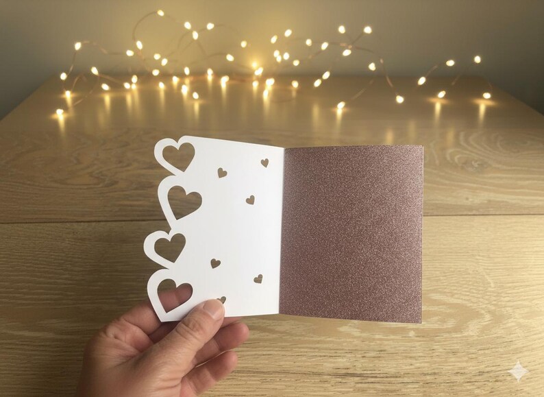 Pu&ograve; includere: Un biglietto di auguri bianco a forma di cuore, con un interno rosa dorato glitterato. Il biglietto presenta una serie di ritagli a forma di cuore e piccoli cuori decorativi. Il biglietto &egrave; aperto, tenuto in mano.