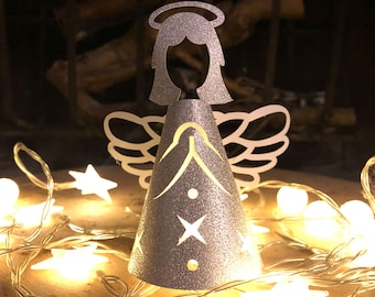 May include: A silver glitter angel decoration with a halo and wings. The angel has a star and other cut-out designs on its cone-shaped body. A string of star-shaped lights and round lights are in the foreground.