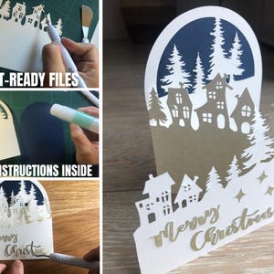 May include: A white paper Christmas card with a layered design featuring a village scene and pine trees. The card has the words "Merry Christmas" in gold script. The card is on a wooden surface.