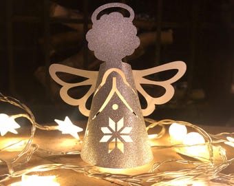 May include: A silver glitter angel Christmas ornament with cut-out snowflake and star designs. The angel has white wings and a halo. A string of star-shaped lights and round lights are in the foreground, creating a warm, festive glow.