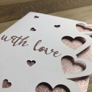 Pu&ograve; includere: Un biglietto bianco con cuori ritagliati e le parole "with love" in corsivo. Il biglietto &egrave; sovrapposto a una superficie scintillante color oro rosa, creando un design romantico ed elegante. Perfetto per un'occasione speciale.