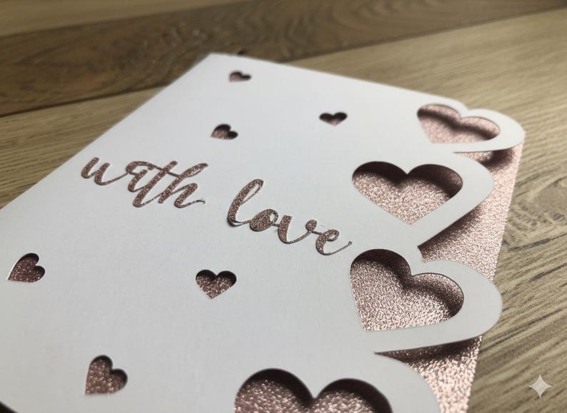 Pu&ograve; includere: Un biglietto bianco con cuori ritagliati e le parole "with love" in corsivo. Il biglietto &egrave; sovrapposto a una superficie scintillante color oro rosa, creando un design romantico ed elegante. Perfetto per un'occasione speciale.