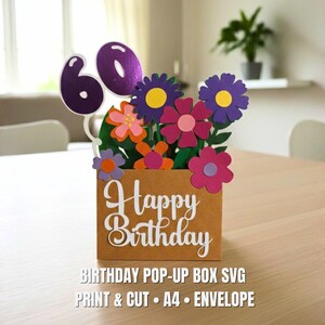 May include: A brown paper pop-up box with the words "Happy Birthday" in white script. The box is filled with colorful paper flowers and the number "60". The image also includes the text "BIRTHDAY POP-UP BOX SVG PRINT & CUT A4 &bull; ENVELOPE".