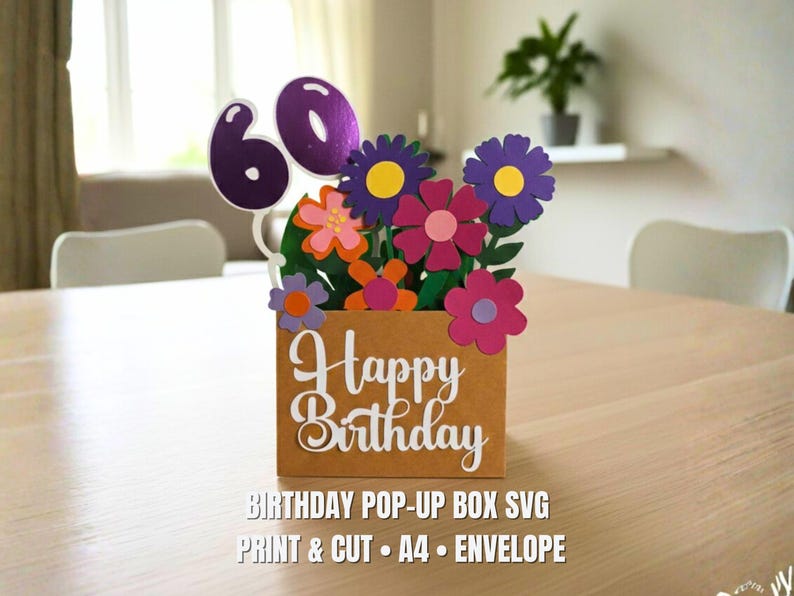 May include: A brown paper pop-up box with the words "Happy Birthday" in white script. The box is filled with colorful paper flowers and the number "60". The image also includes the text "BIRTHDAY POP-UP BOX SVG PRINT & CUT A4 &bull; ENVELOPE".