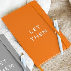 May include: Orange notebook with the words "LET THEM" in white. A light blue pen rests on the notebook. A gray notebook with the same text is partially visible. The notebooks have elastic bands.