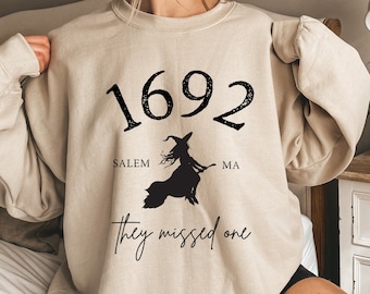May include: A beige sweatshirt with a black graphic print that says "1692 Salem MA they missed one" with a silhouette of a witch flying on a broomstick.