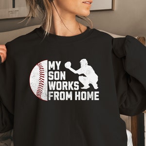 May include: Black sweatshirt with a white graphic of a baseball and a baseball player catching a ball. The text on the graphic reads "My son works from home."