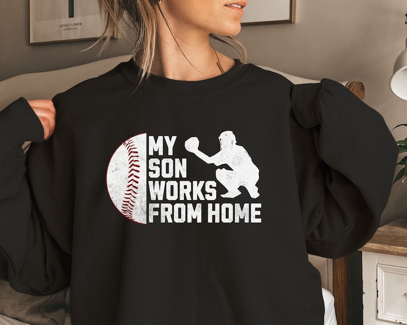 May include: Black sweatshirt with a white graphic of a baseball and a baseball player catching a ball. The text on the graphic reads "My son works from home."