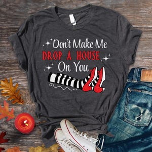 May include: A dark gray t-shirt with the text "Don't Make Me Drop A House On You" and a graphic of a witch's legs wearing red shoes with black and white stripes.