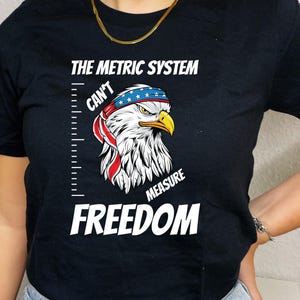 May include: Black t-shirt with a white graphic of an eagle wearing a red, white, and blue bandana. The graphic says "The Metric System Can't Measure Freedom".