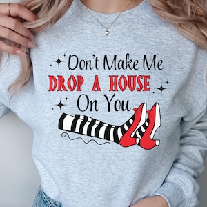 Op de afbeelding: Grijs sweatshirt met de tekst "Don't Make Me Drop A House On You" in rood en zwart. De afbeelding toont een paar rode schoenen met zwart-witte strepen.