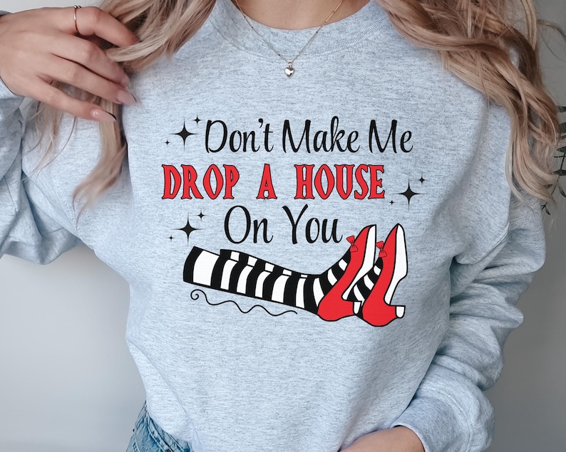 Op de afbeelding: Grijs sweatshirt met de tekst "Don't Make Me Drop A House On You" in rood en zwart. De afbeelding toont een paar rode schoenen met zwart-witte strepen.