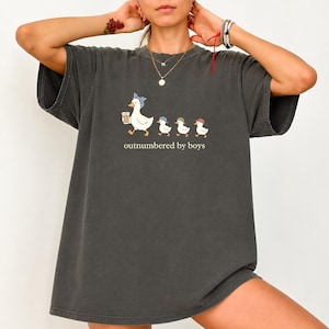 May include: A charcoal gray short-sleeve t-shirt featuring a graphic of a mother duck wearing a blue polka dot headscarf holding a coffee cup, followed by three small ducklings in colorful baseball caps, with the text outnumbered by boys.